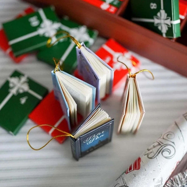 Mini book ornaments with gold hanging strings beside red and green boxes